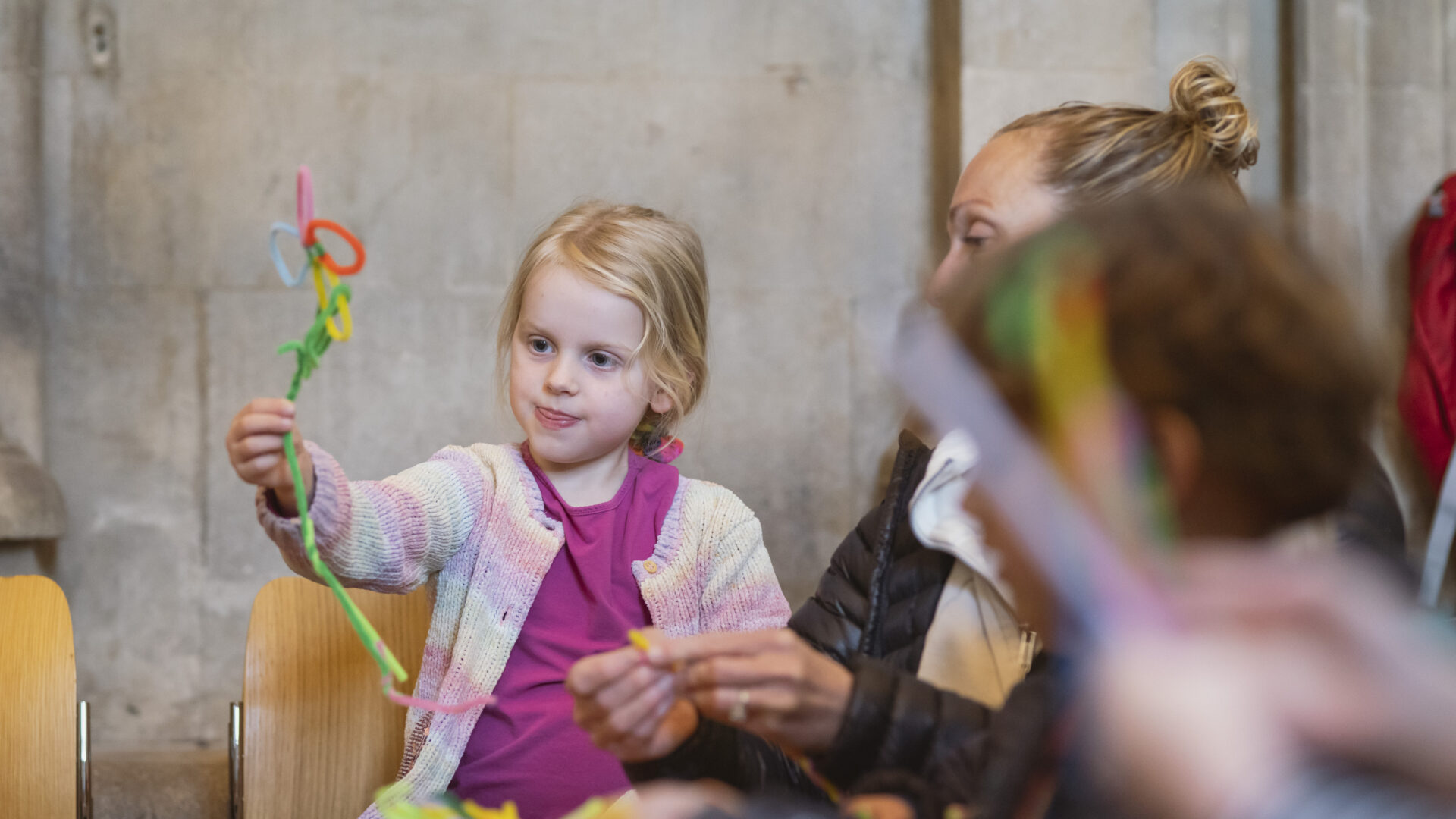 Little girl holding up a pipe cleaner flower, sat beside her mum at Crafty Cathedral